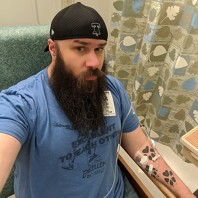 Man with long beard and backwards cap sitting with an IV in his tattooed arm taking a selfie.