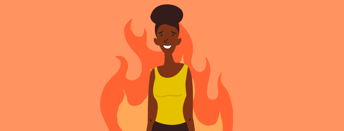 A woman is standing as flames burn behind her. She has a big smile and sweat running down her face.